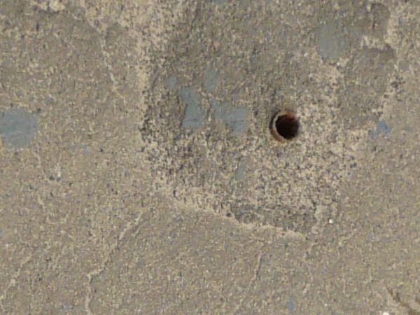 Road edge texture with a small strip of sidewalk with small blue marks on it and small metal holes in it. The sidewalk runs between a patch of brown weeds at the bottom, and rough, grey road at the top.